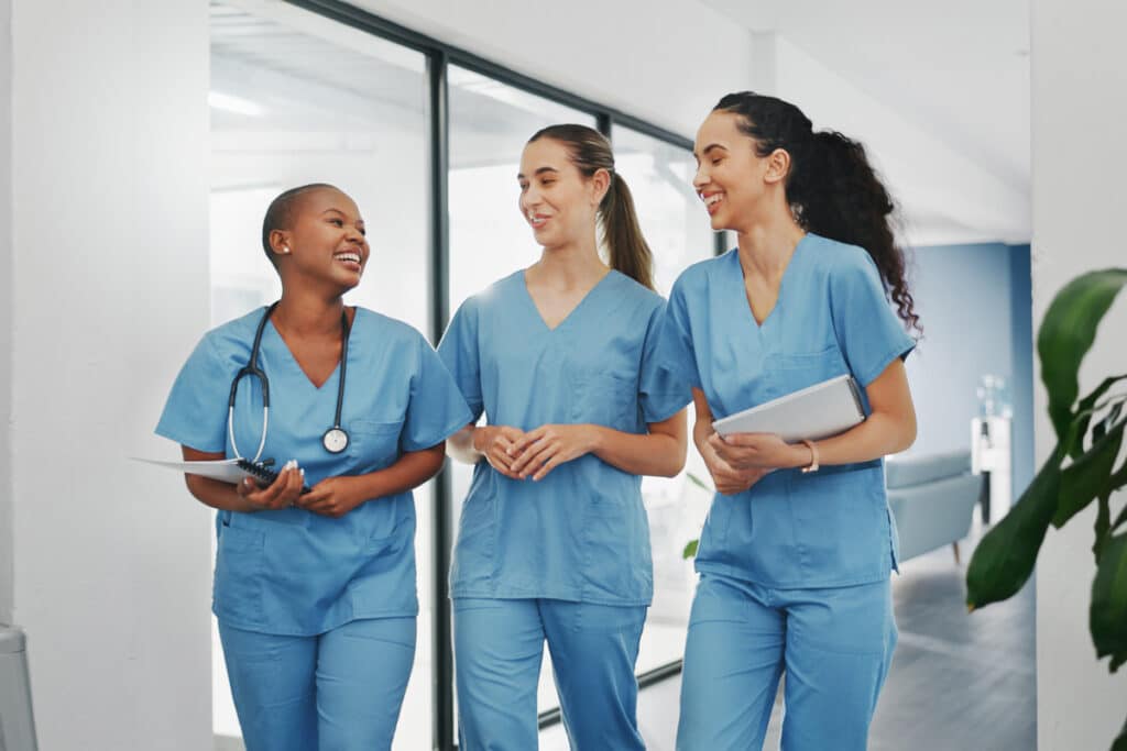 Three nurses in blue scrubs walk down a hospital corridor, smiling and chatting. One holds a stethoscope.